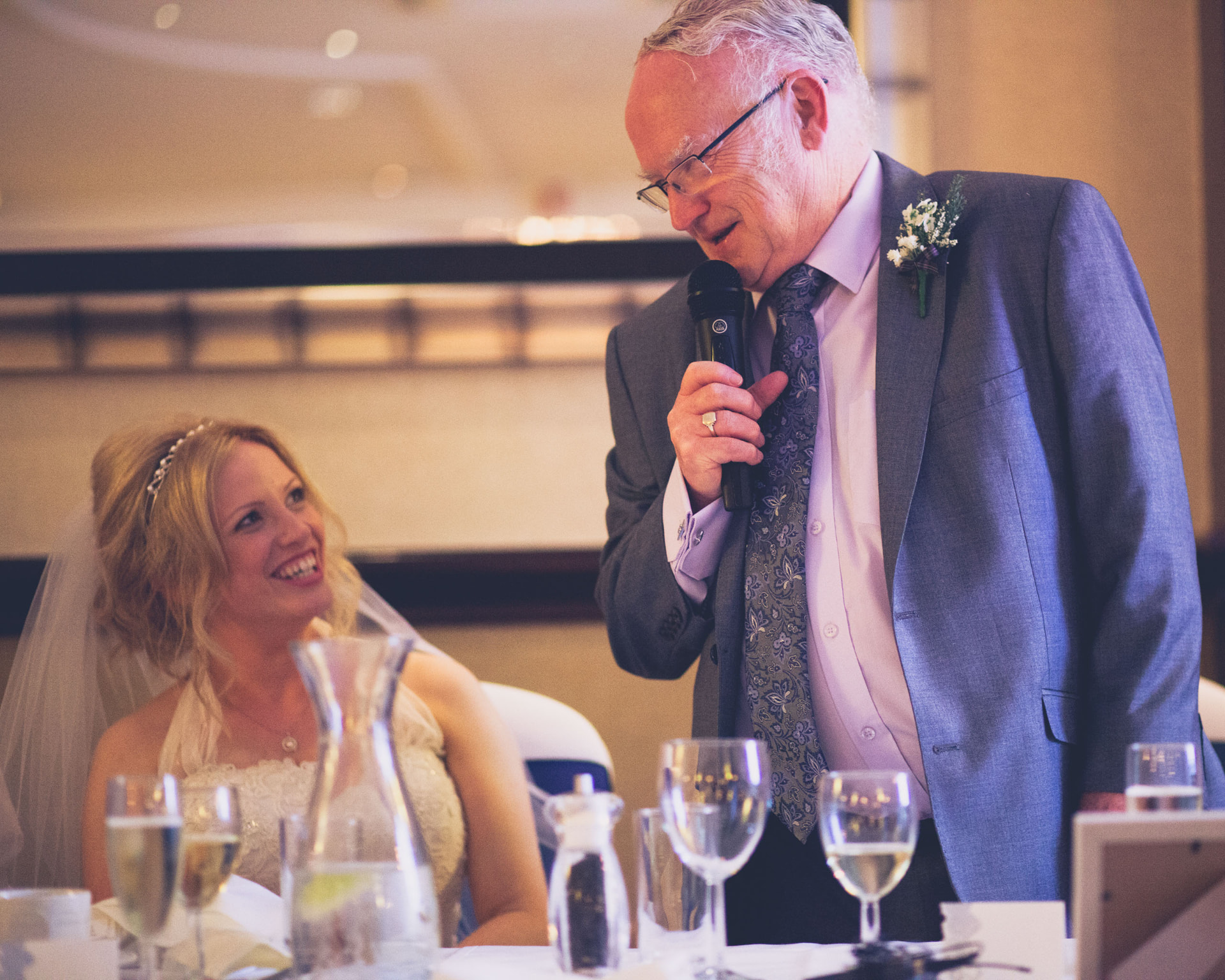 Bride laughing as her father gives his speech with a microphone at the wedding reception