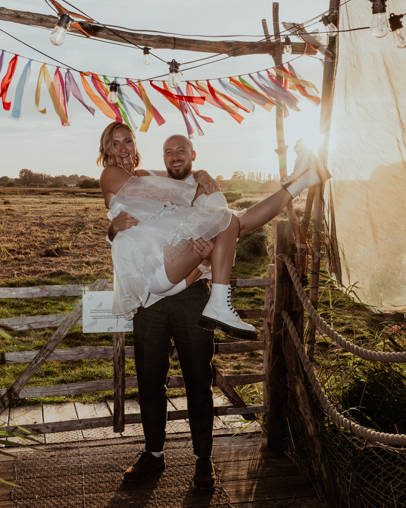 Groom lifting his bride at golden hour beneath ribbon bunting and festoon lights