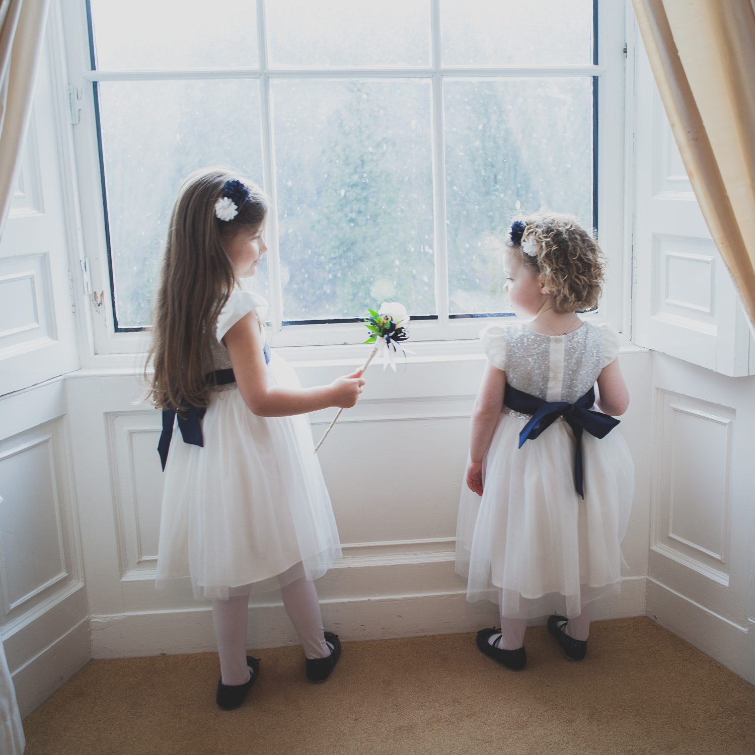 Two flower girls in ivory dresses looking out of a rainy window before the ceremony